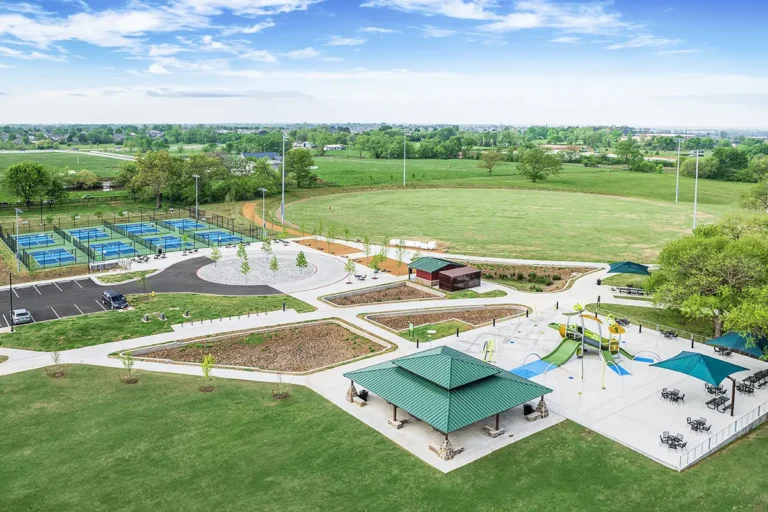 Aerial view of a park showing tennis courts, a circular plaza, playgrounds with slides, sunshades, and green fields under a clear blue sky.