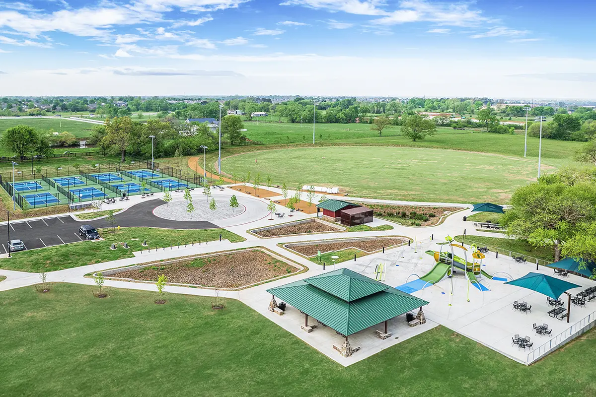 Aerial view of a park showing tennis courts, a circular plaza, playgrounds with slides, sunshades, and green fields under a clear blue sky.