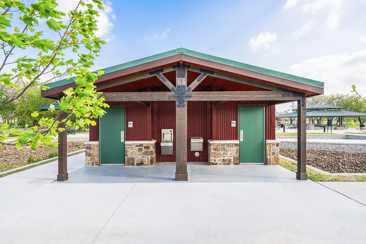 A park restroom with green doors, brown siding, and stone accents, under a clear blue sky. Nearby trees and open pavilion create a welcoming park feel.