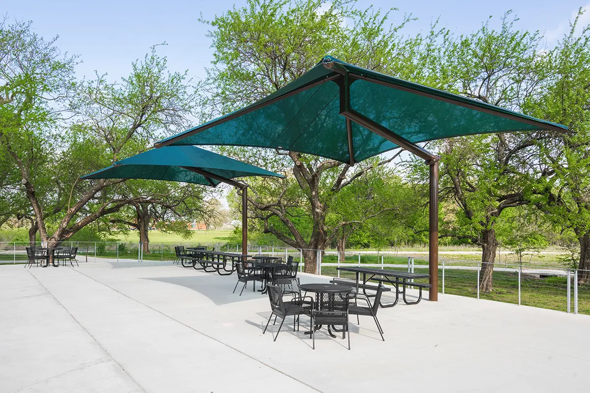 Outdoor patio with teal shade sails overhead. Black tables and chairs are arranged on a concrete surface. Surrounded by lush green trees, conveying a peaceful and inviting atmosphere.