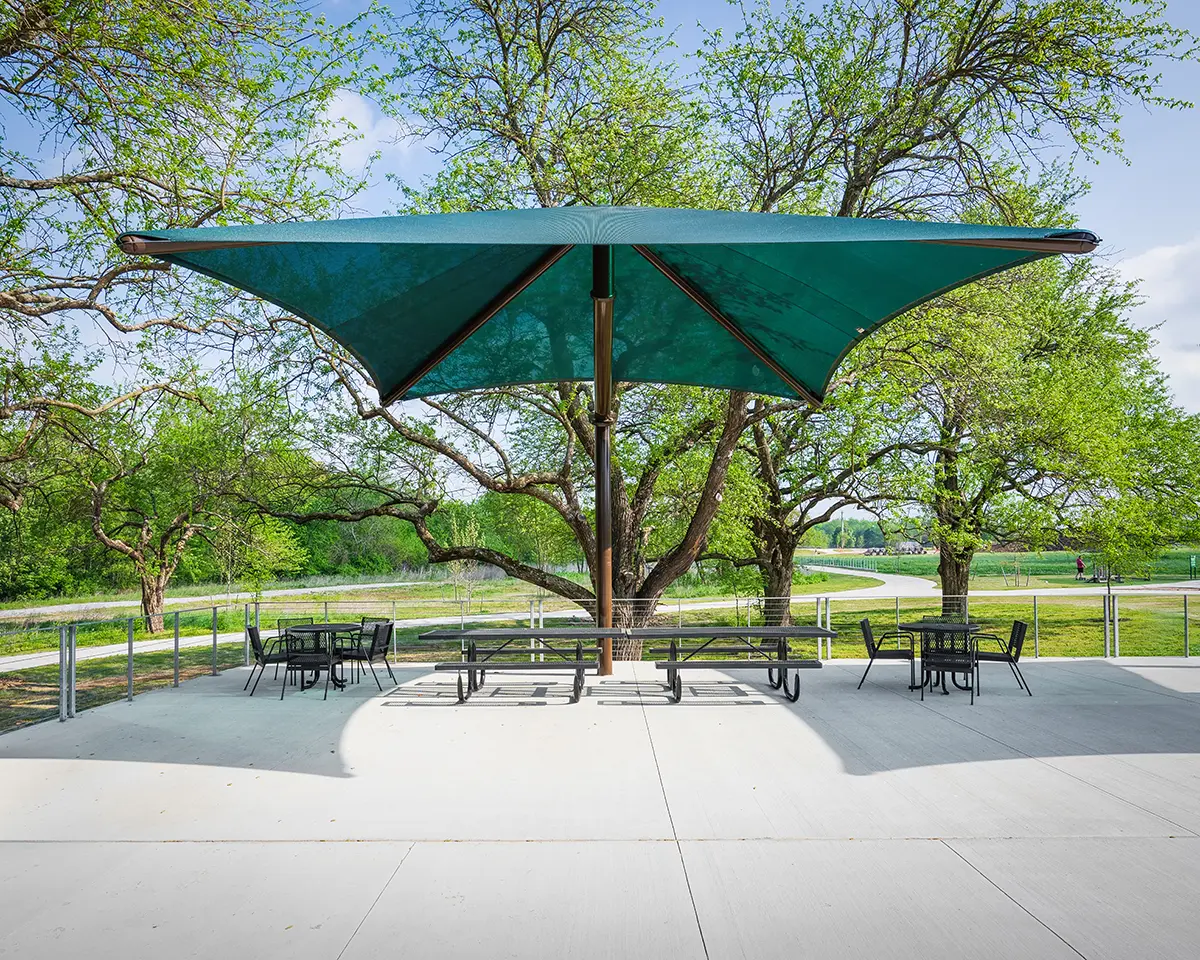 A large green umbrella shades picnic tables on a sunny patio, surrounded by lush trees and a winding path, creating a serene outdoor setting.