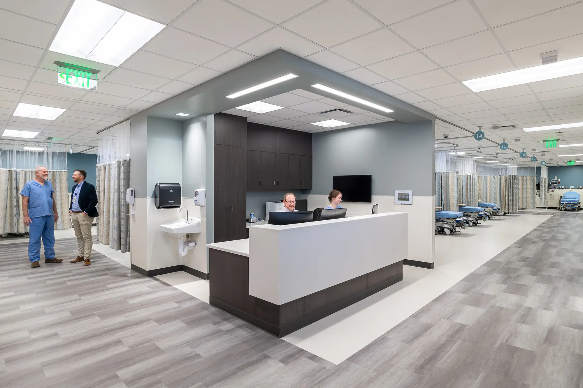 A modern hospital reception area with two staff at a desk, open patient beds with privacy curtains in the background, and two men conversing on the left.