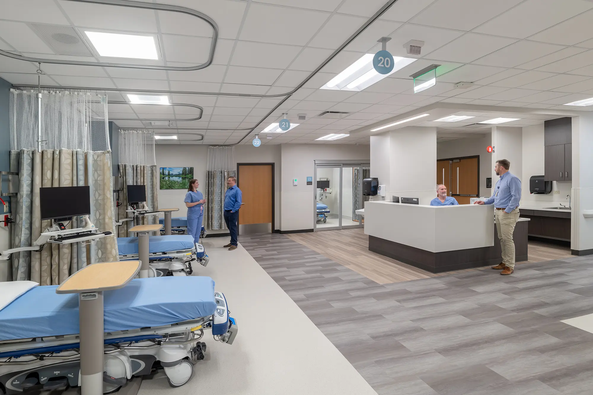 A hospital ward with empty beds and monitors on the left. Two people converse by the door, while two others interact at a central nurses' station. Clinical setting.