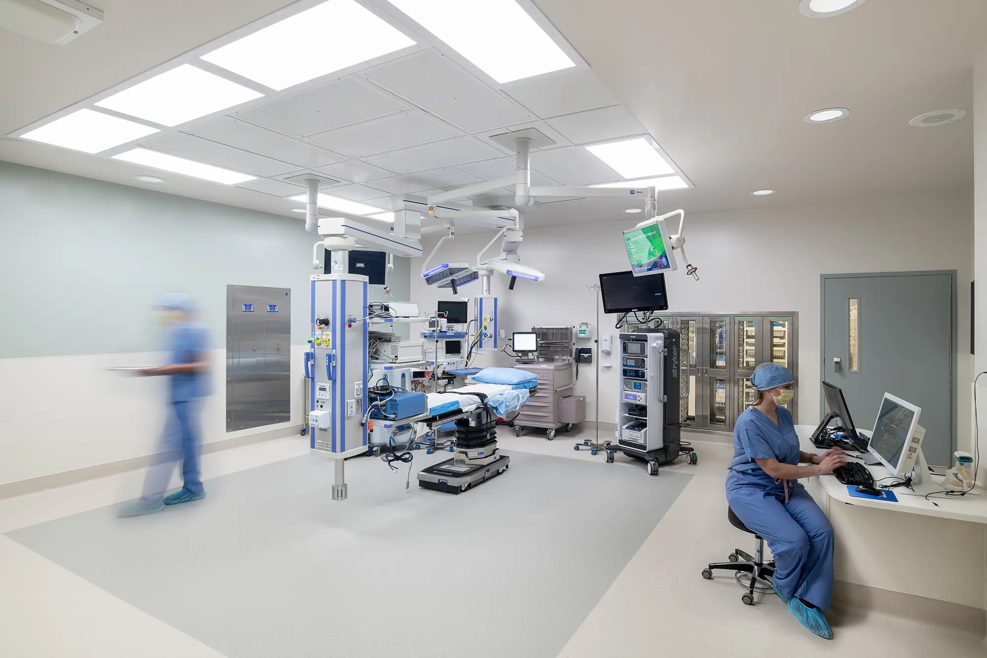 Modern operating room with advanced medical equipment, bright lighting, and two medical staff in blue scrubs; one seated at a computer, the other blurred walking.