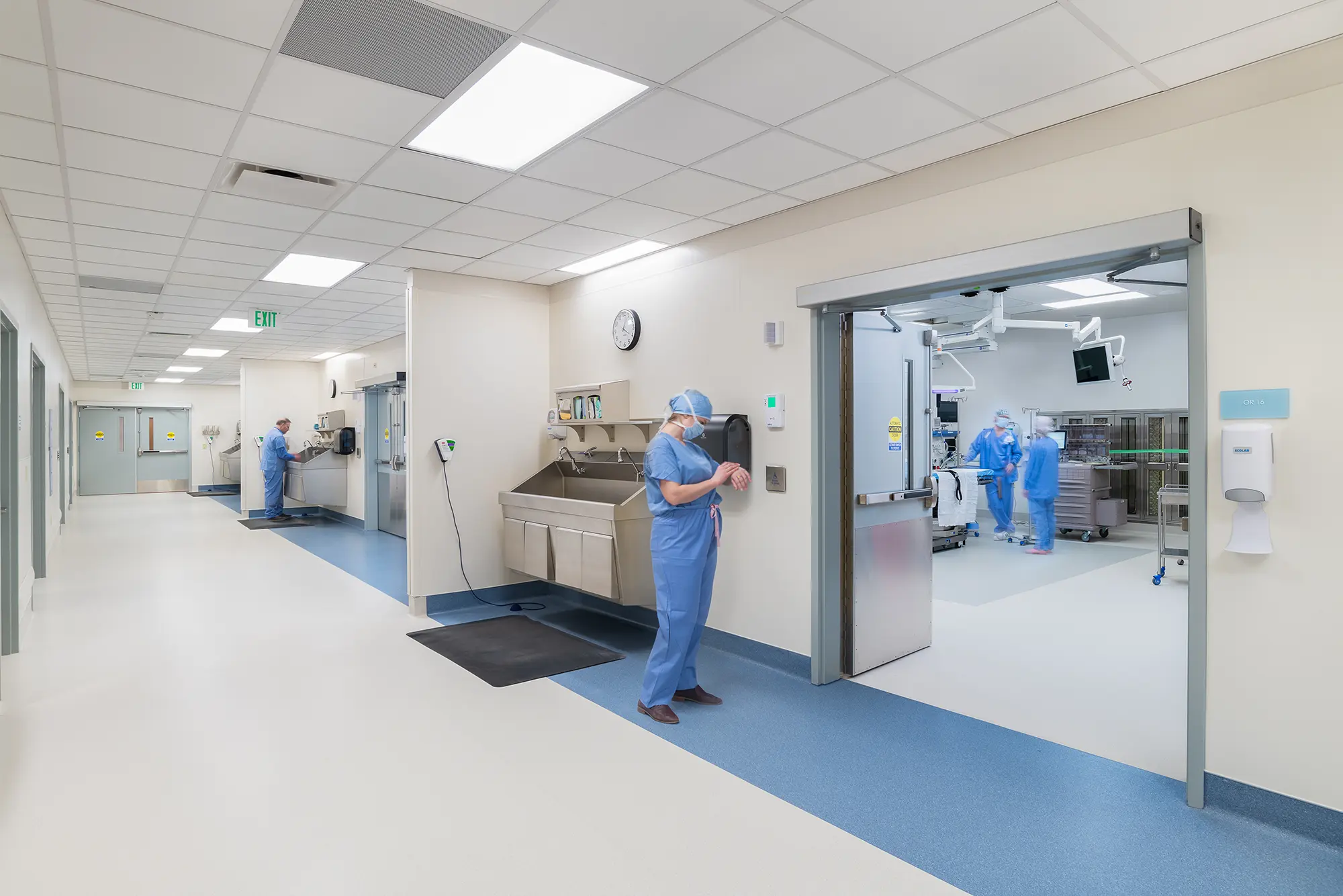 Hospital corridor with doctors in scrubs and masks, preparing outside an operating room. The area is clean and brightly lit, reflecting a clinical atmosphere.