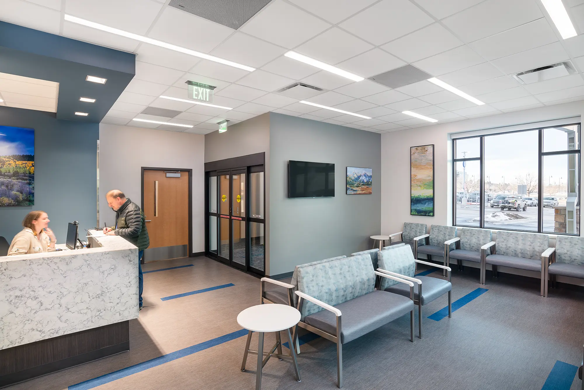 A modern waiting room with gray seating and a check-in desk. A person talks to a receptionist. Large windows offer natural light, with paintings on the walls.