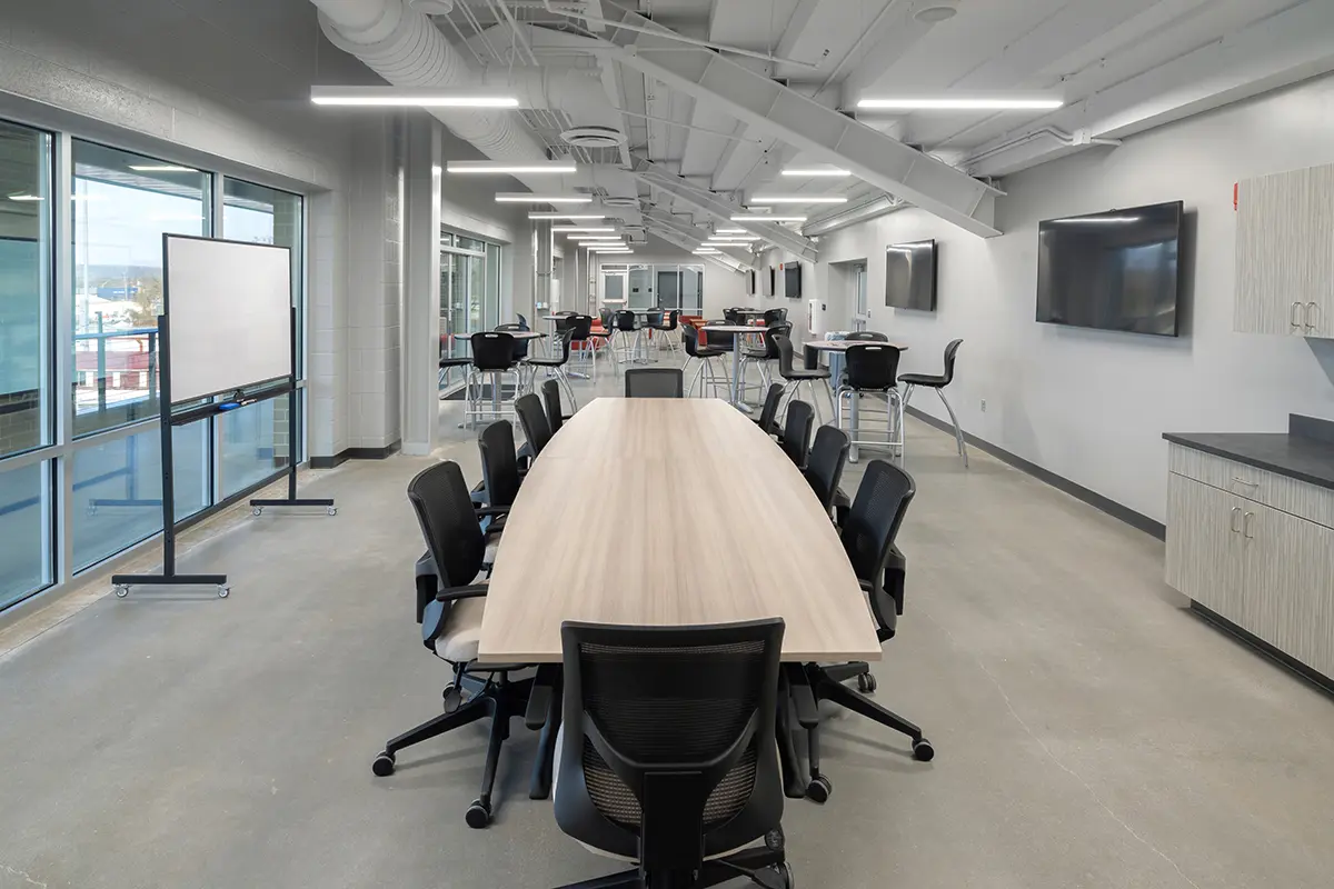 Modern conference room with a long wooden table surrounded by black chairs. Bright lighting, large windows, whiteboard, and wall-mounted screens create a professional atmosphere.