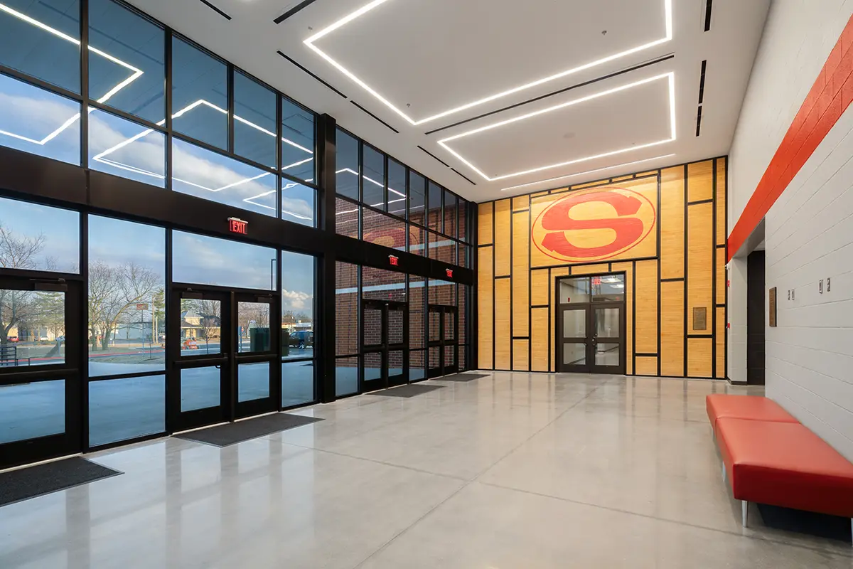 Modern lobby with large glass windows, geometric ceiling lights, and a bold red "S" logo on a wooden accent wall. A red bench sits on the right.