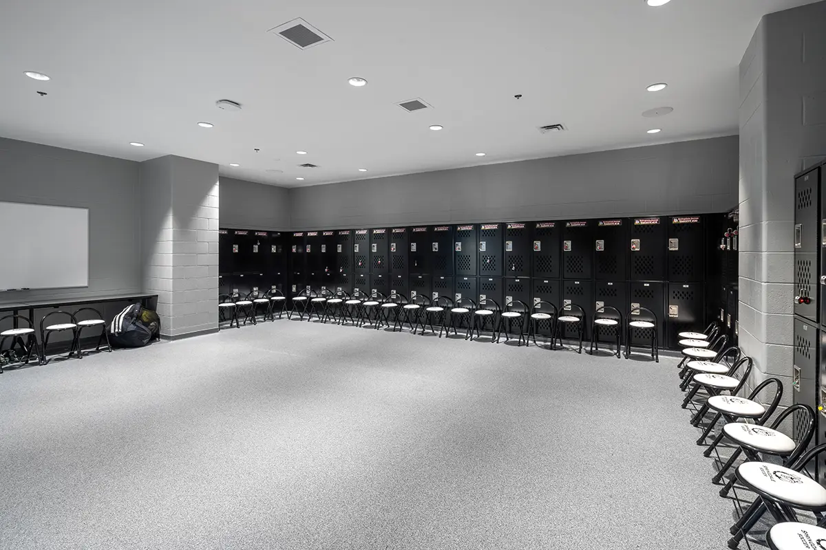 Spacious locker room with rows of black lockers along two walls, each flanked by folding chairs. Bright lighting and a calm, organized ambiance.
