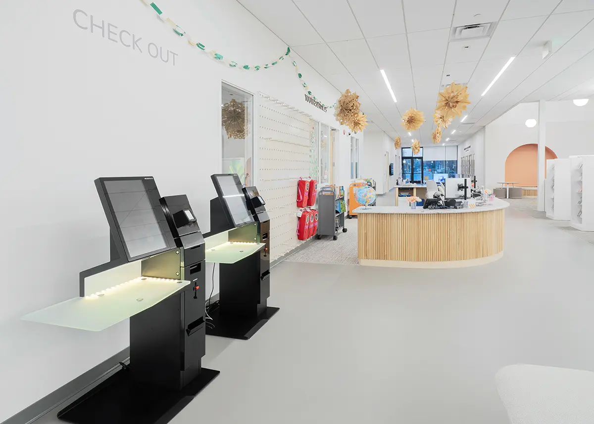 Bright library checkout area with self-service kiosks, a wooden desk, and playful hanging decorations, conveying a modern and inviting atmosphere.