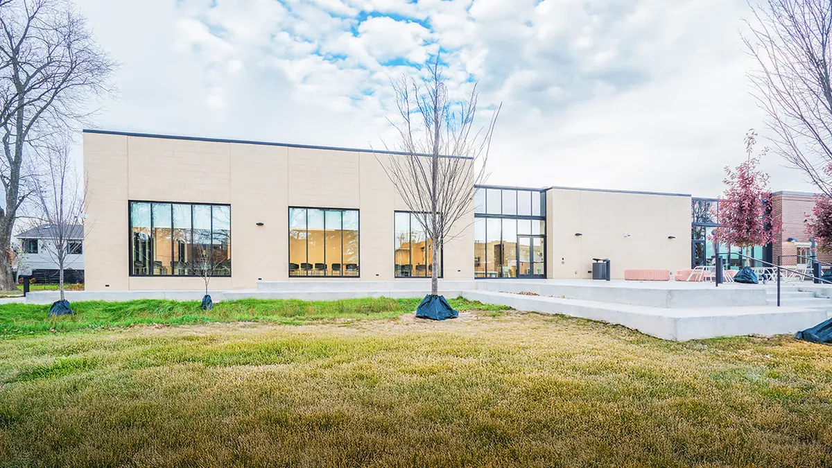 Modern building with large windows on a bright day, surrounded by a grassy field and bare trees. The sky is cloudy, conveying a serene atmosphere.