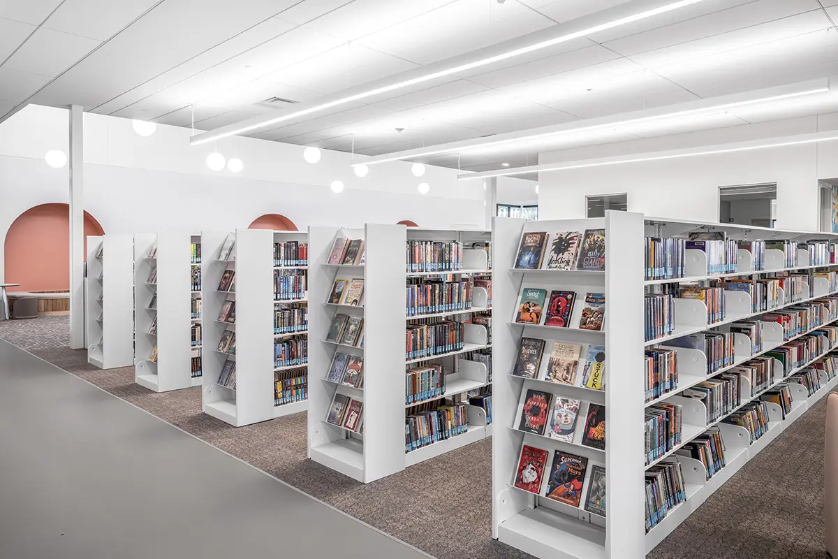 Modern library interior with white bookshelves filled with books and graphic novels. Soft lighting and muted peach wall accents create a calm atmosphere.