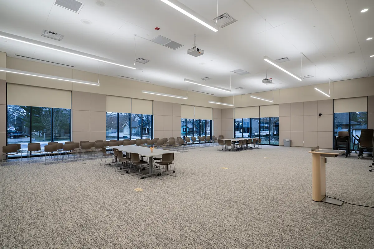 Spacious conference room with beige walls and large windows showing trees outside. Rows of chairs and tables, with a podium in the foreground.