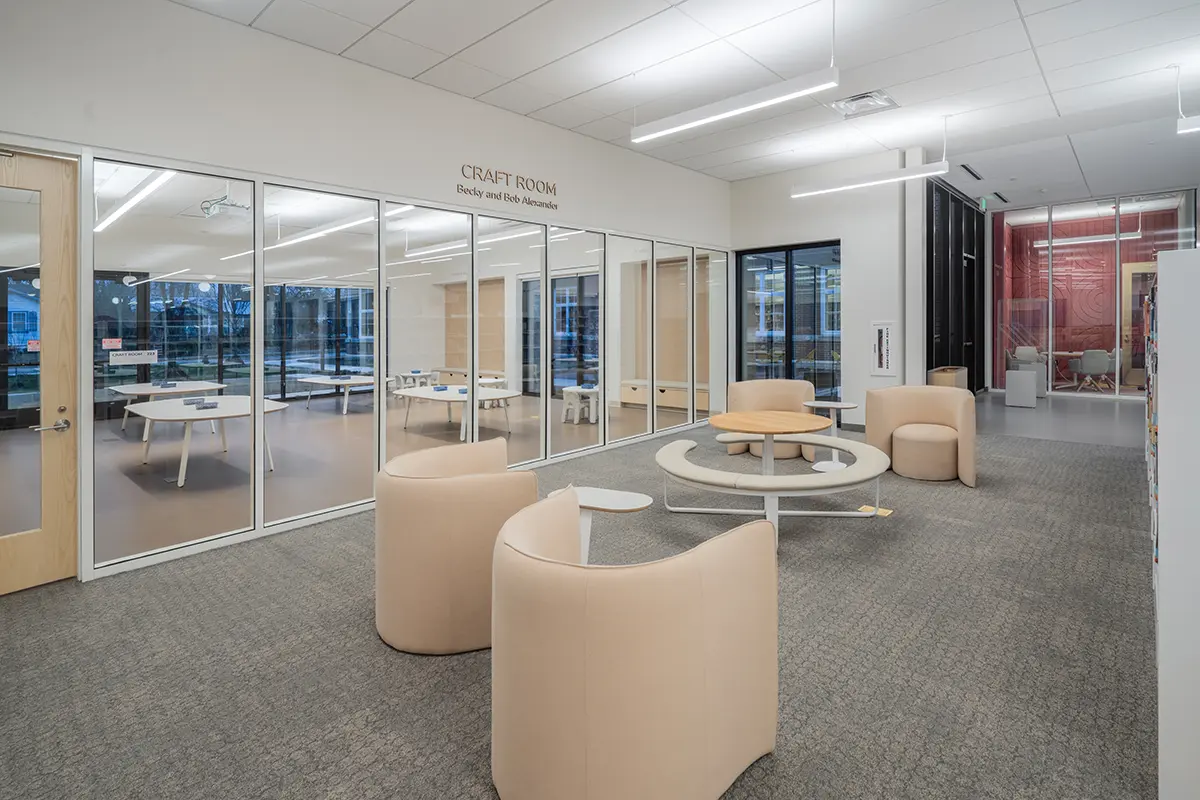 A modern craft room with glass walls, beige seating, round tables, and ample natural light. The space feels open and inviting, ideal for creative activities.
