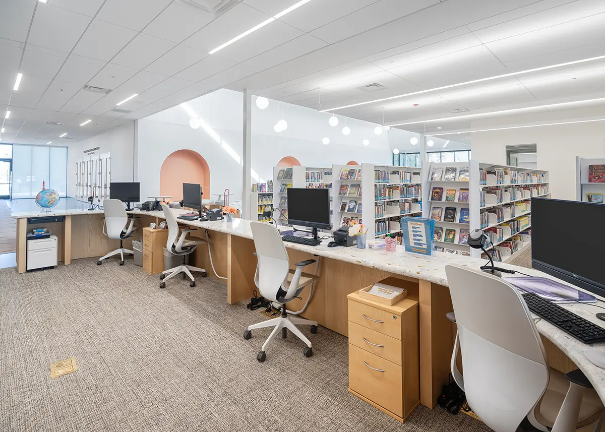 Modern library with sleek desk stations, computers, and swivel chairs. Shelves filled with colorful books in a bright, open space with soft lighting.