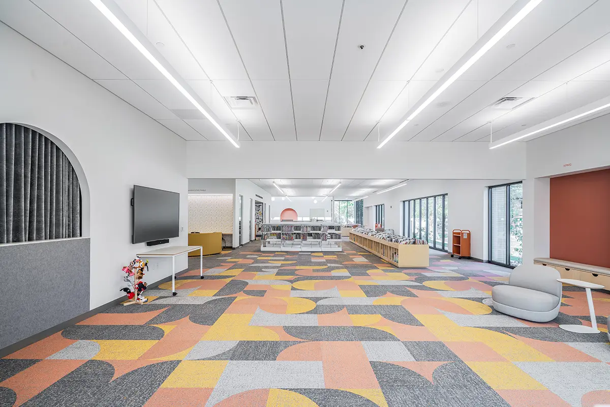 Modern library interior with colorful geometric carpeting in orange, gray, and yellow tones. Bright lighting, large windows, comfy seating, and a TV.