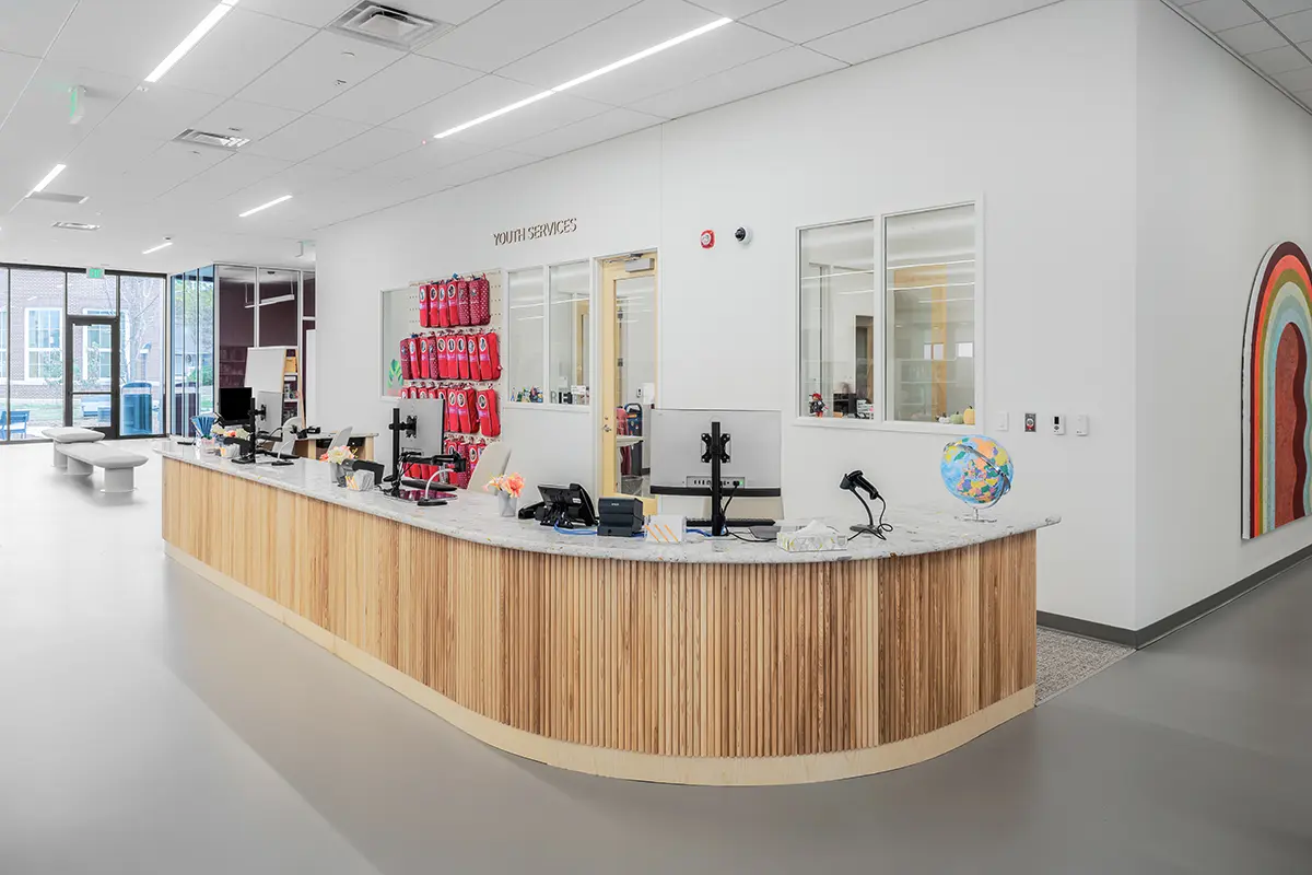 A modern youth services library desk with a curved wooden front, decorated with a globe and office supplies. Bright, open space with a rainbow mural.
