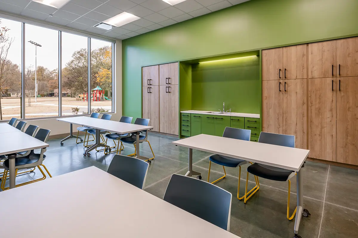 A brightly lit classroom with green walls, wooden cabinets, and tables arranged neatly. Large windows show an outdoor playground, creating an inviting atmosphere.