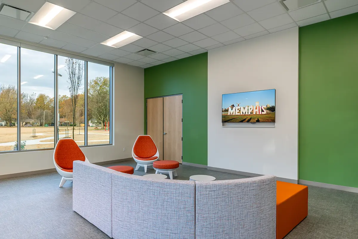 Modern waiting room with gray sofa and orange chairs facing a TV displaying "Memphis." Large window and green accent wall create a bright, inviting atmosphere.