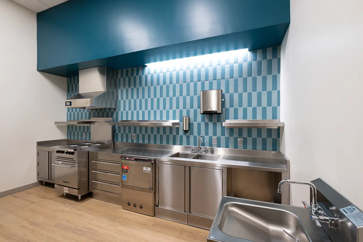 Modern commercial kitchen with stainless steel appliances, blue tiled backsplash, and wooden floors. Well-lit, clean, and organized space.