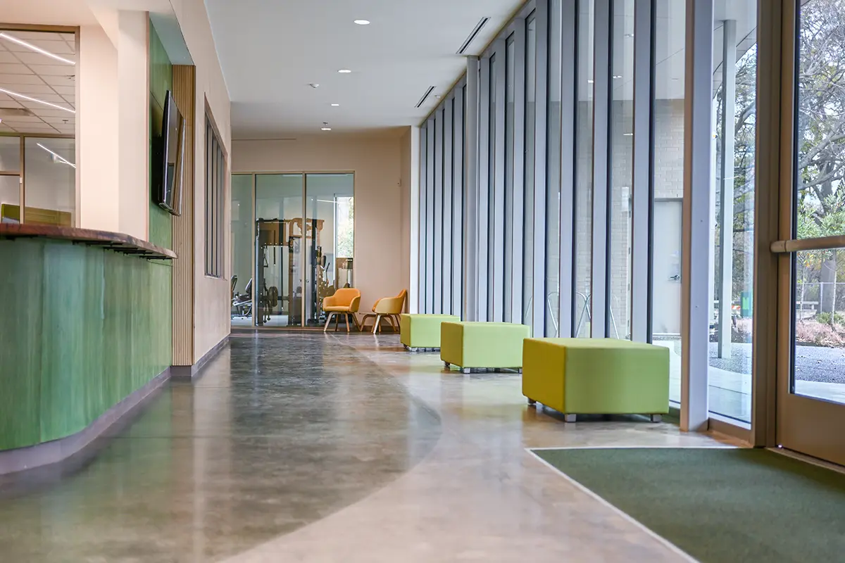 Modern hallway with floor-to-ceiling windows, green and beige seating, and a sleek reception desk. Warm lighting, open space, and a calm ambiance.