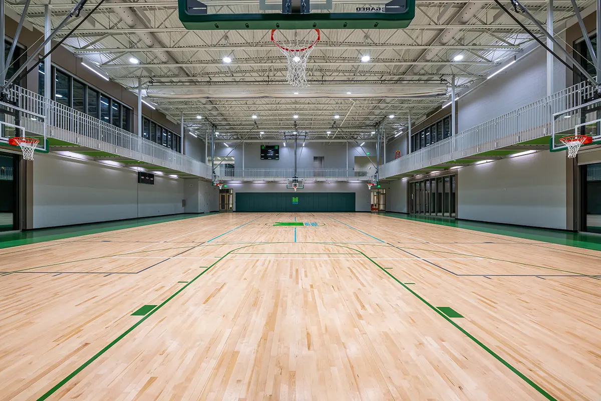 Empty indoor basketball court with polished wooden floors, green markings, and multiple hoops. The space is well-lit, spacious, and clean.