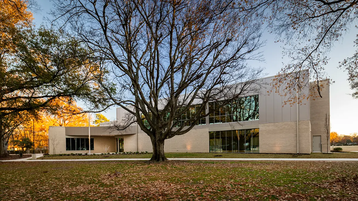 A modern two-story building with large windows is framed by a bare tree, set against a backdrop of colorful autumn foliage and a clear sky.