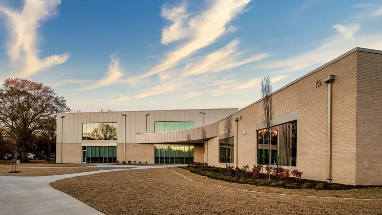 Modern, single-story brick building with large windows under a vibrant blue sky with wispy clouds. It is surrounded by neatly trimmed grass and a few trees.