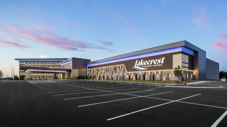 Modern casino hotel exterior at sunset, with a large, empty parking lot in front. The building features sleek lines, illuminated signage, and a serene sky.
