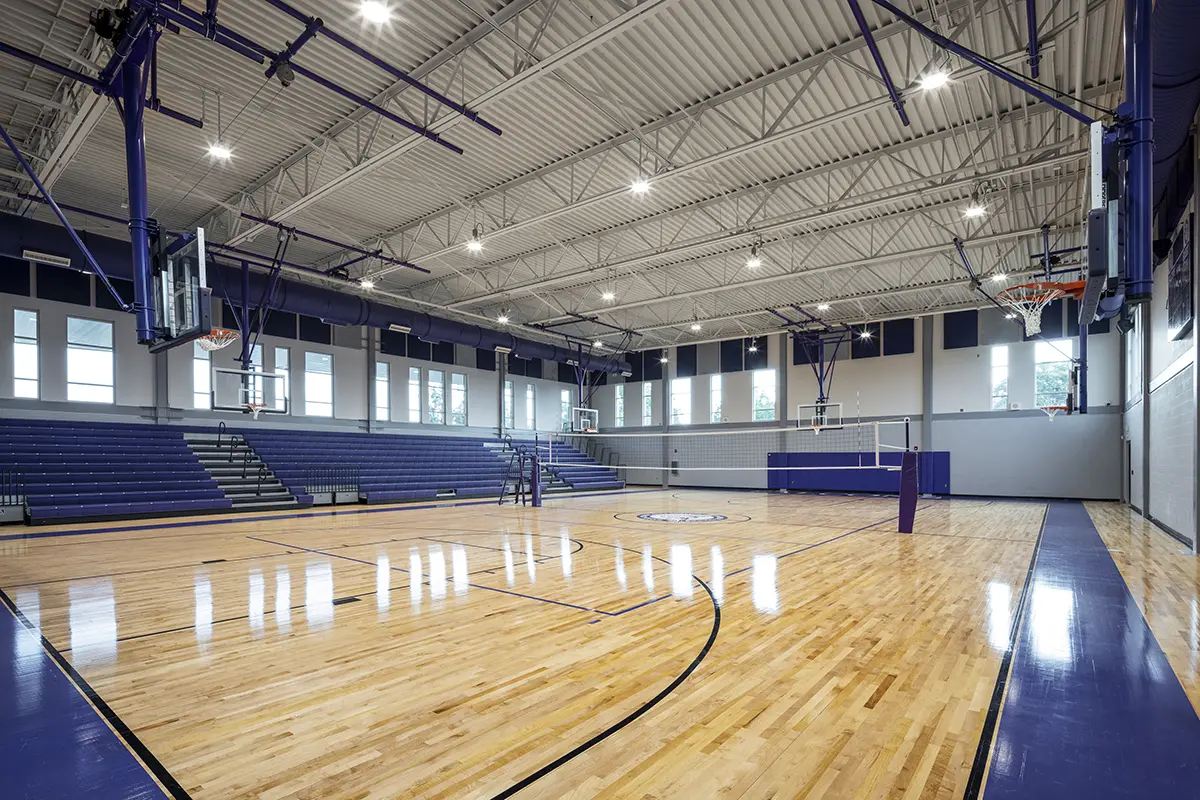 Bright, spacious gymnasium with polished wood flooring, equipped for volleyball and basketball. Empty bleachers line the side; windows and lighting create an airy feel.