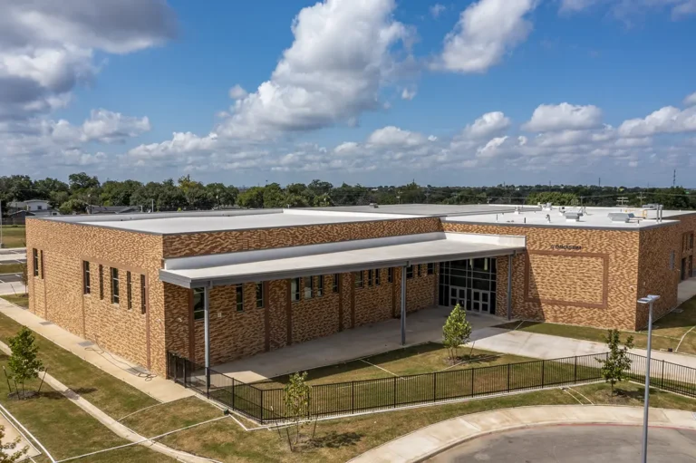 A modern, single-story brick building with large windows, seen on a sunny day. It features a covered entrance shaded by a roof extension, surrounded by a fence and young trees.