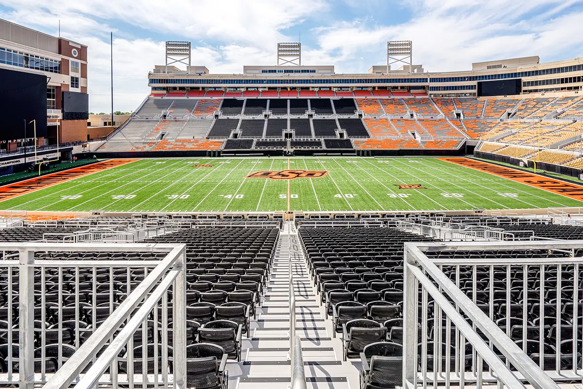 Empty football stadium with a green field marked with white lines and an orange logo at the center. Orange and black seating surround the field.
