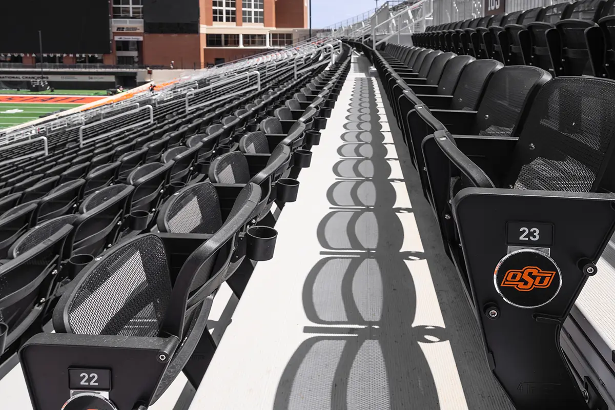 Stadium seats with black mesh backs and OSU logo form uniform rows under bright sunlight. Shadows create a rhythmic pattern along the concrete aisle.