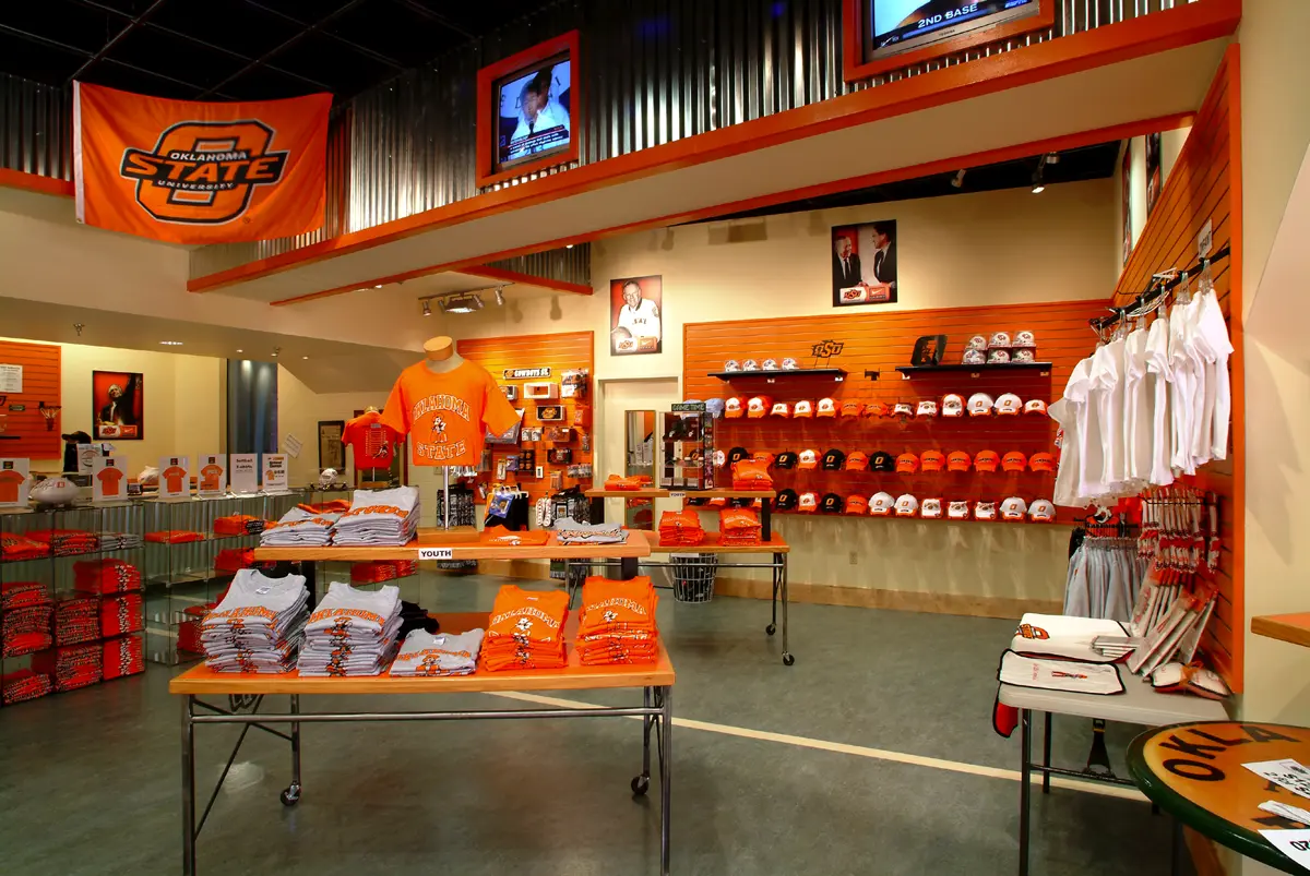 Brightly lit store interior showcasing Oklahoma State merchandise. Orange T-shirts, caps, and sportswear are neatly displayed on tables and shelves. Vibrant team spirit.