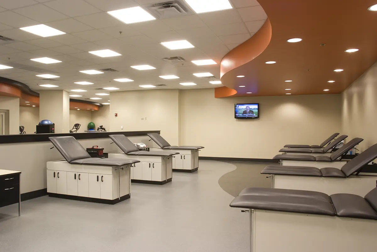 A spacious, modern physical therapy clinic features multiple padded exam tables, a curved orange ceiling, bright lighting, and a wall-mounted TV.