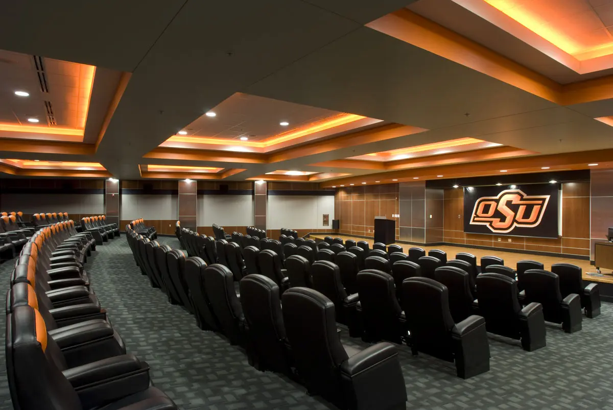 Lecture hall with tiered leather seating and orange-lit ceiling. A stage features a large logo against wooden paneling. Modern and professional ambiance.