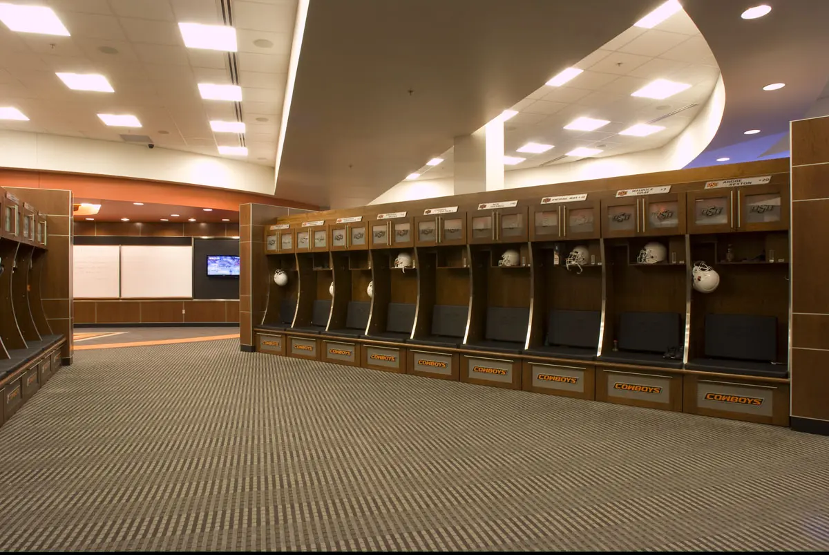 Spacious football locker room with wooden lockers, helmets, and comfortable seating. Bright overhead lights create a clean, organized atmosphere.