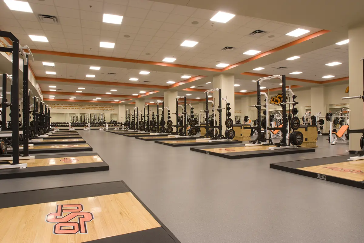 Spacious gym with rows of squat racks and weights on wood platforms. Bright overhead lighting highlights the organized, clean, and modern facility.
