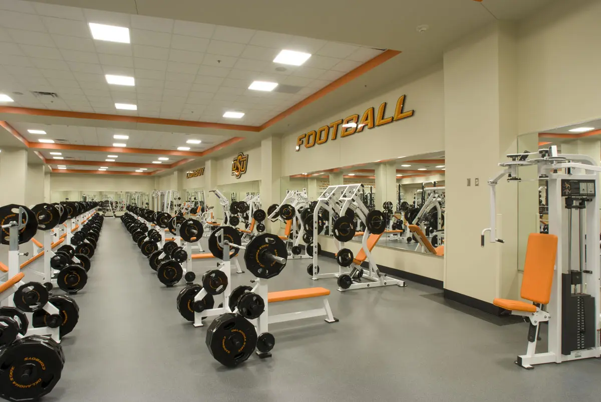 Modern gym with rows of dumbbells, benches, and weight machines, featuring orange accents. "Football" sign above mirrors, conveying a sporty tone.