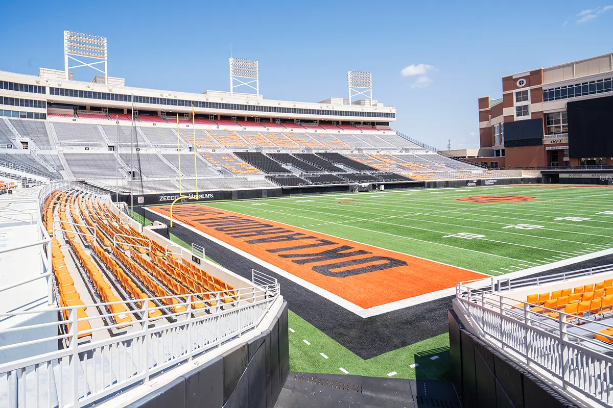 Empty football stadium in bright sunlight, featuring rows of orange and black seats. Green field marked with "OKLAHOMA" in end zone, and a clear blue sky above.