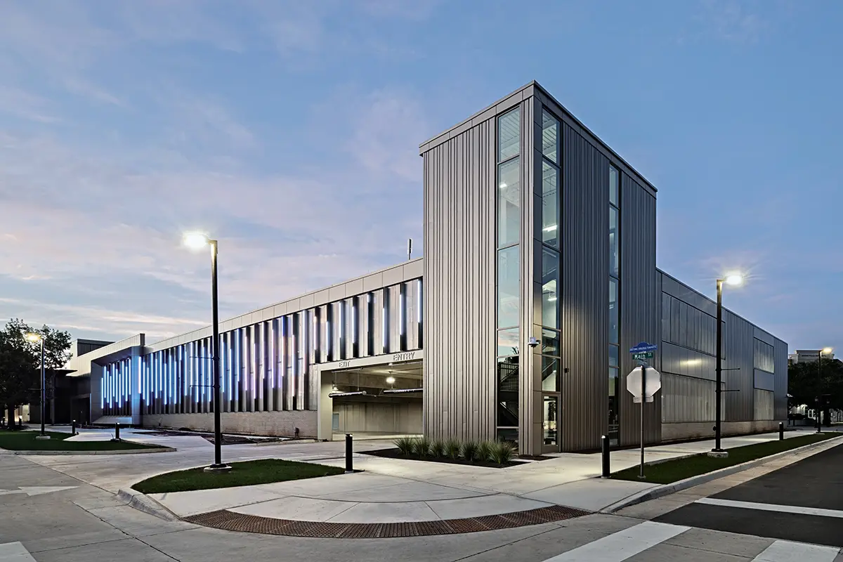 Modern building with large glass windows and vertical metal panels, illuminated by street lamps at dusk. The sky is a gradient of pink and blue.
