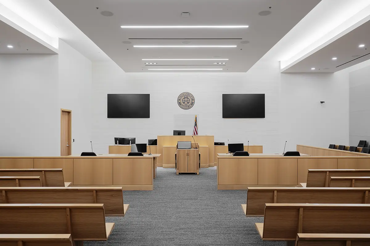 Modern courtroom with wooden benches and desks, two large screens, and an American flag behind the judge's bench. Bright, orderly, and formal atmosphere.