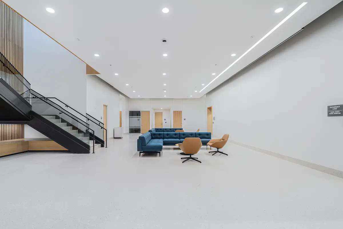 Spacious, modern lobby with white walls and floor, featuring a large blue sectional sofa and two mustard swivel chairs. Nearby, a sleek black staircase ascends.