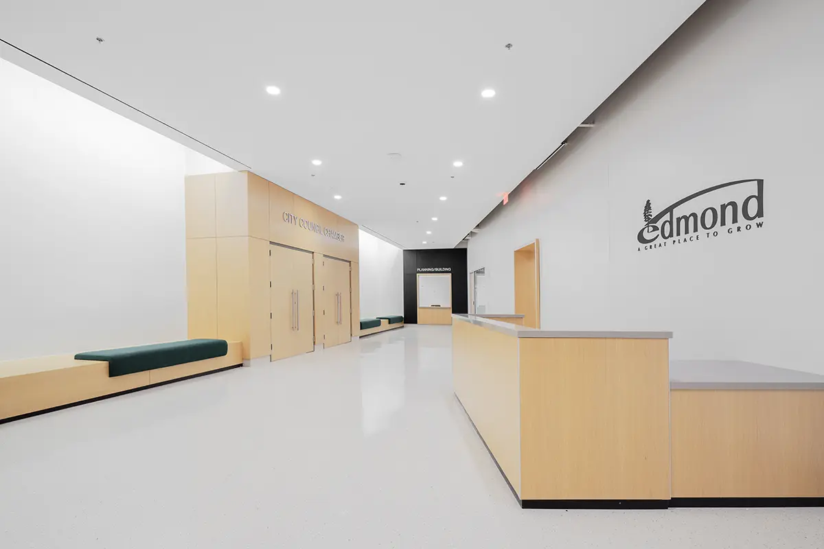 Minimalist hallway with light wood paneling, recessed lighting, and green cushioned benches. "Edmond" logo and "City Council Chambers" sign visible.