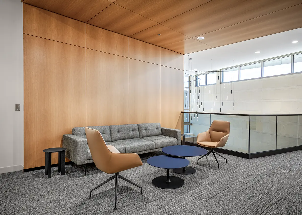 Modern lounge area with a gray sofa, two tan chairs, a black side table, and two round blue coffee tables. The space is bright with wooden walls.