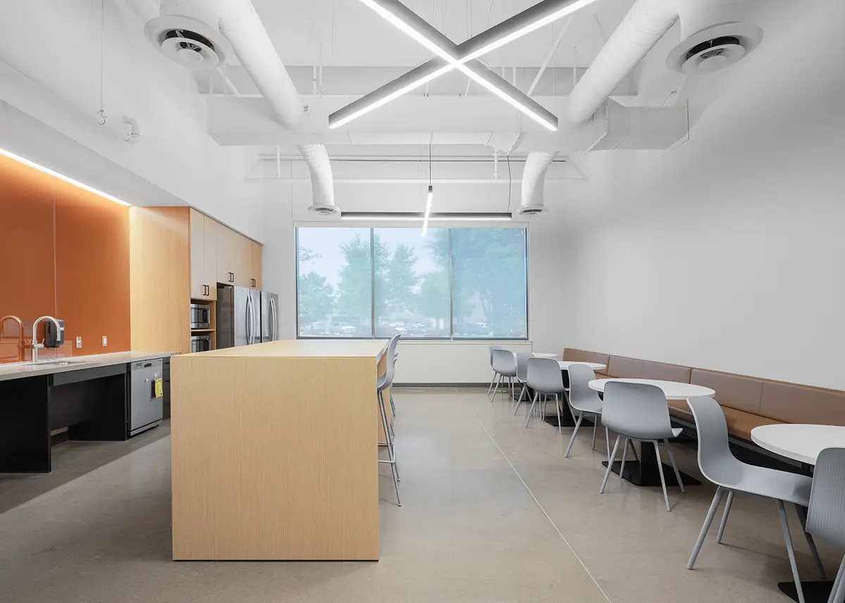 Modern office kitchen with sleek design elements. Features a wooden island, round tables with gray chairs, large window, and geometric ceiling lights.
