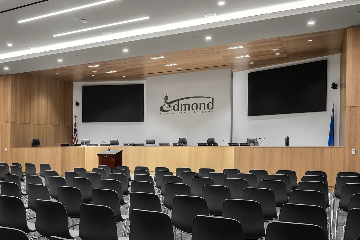 Modern conference room with wooden accents and rows of empty black chairs facing a podium. Two large screens and "Edmond: A Great Place to Grow" sign visible.