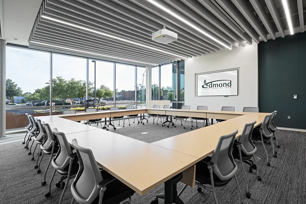 Bright conference room with U-shaped table setup, gray chairs, and large windows showcasing a view of trees. "Edmond" logo is displayed on a wall screen.