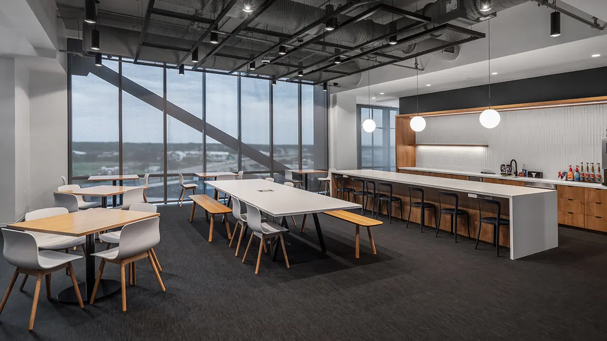 Modern break room with natural light, featuring long tables, white chairs, wooden benches, and a kitchen area with bar stools. Large windows create an airy feel.