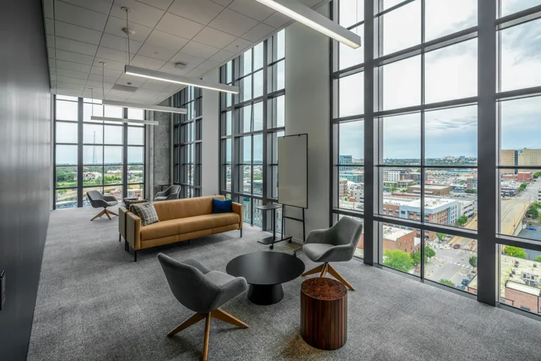 Modern office lounge with large windows and city view. Features a brown sofa, gray chairs, round black table, and sleek lighting, creating a calming atmosphere.